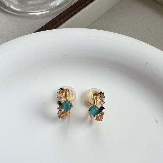 Close-up of dainty 18k gold double hoop clip-on earrings featuring one band of clear pavé diamonds and one band with a single sapphire blue gemstone.