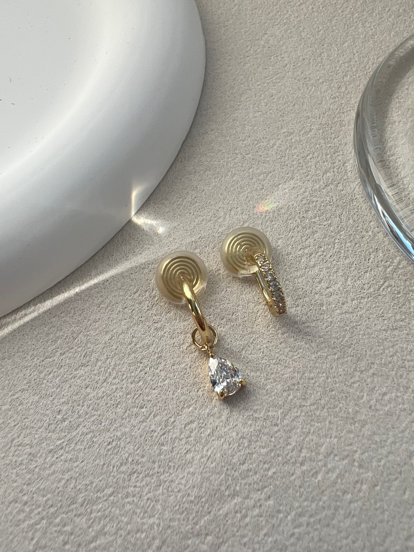 A matching asymmetrical set of two diamond clip-on earrings. One is a classic diamond pave hoop, and the other is a pave hoop with a dangling diamond drop. Both feature clear, invisible resin clips for non-pierced ears