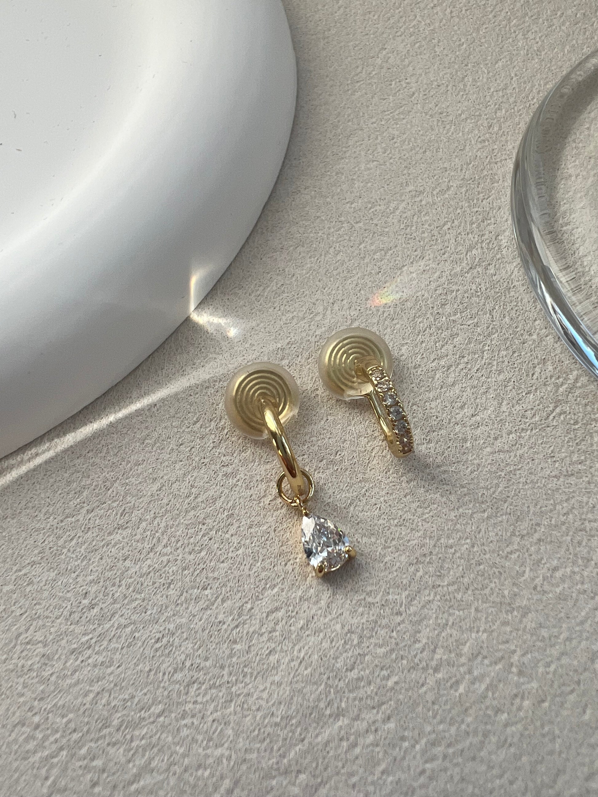 A matching asymmetrical set of two diamond clip-on earrings. One is a classic diamond pave hoop, and the other is a pave hoop with a dangling diamond drop. Both feature clear, invisible resin clips for non-pierced ears