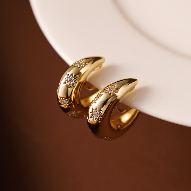 A pair of Earclipse chunky gold dome clip-on hoop earrings featuring engraved starburst patterns with sparkling crystal centres, resting on the edge of a white ceramic plate
