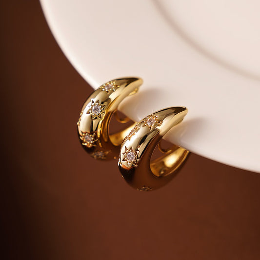 A pair of Earclipse chunky gold dome clip-on hoop earrings featuring engraved starburst patterns with sparkling crystal centres, resting on the edge of a white ceramic plate