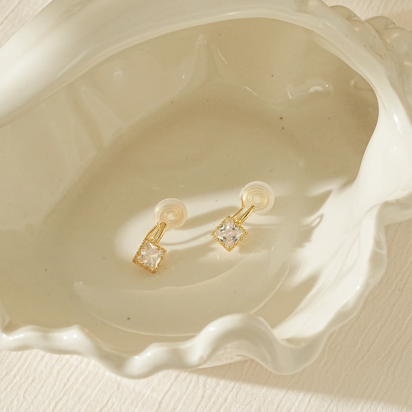 A pair of gold princess cut clip-on earrings laid flat against a soft, creamy background.