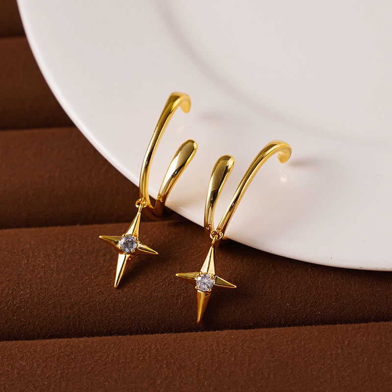 A pair of Earclipse gold illusion clip-on earrings featuring a double-band cuff design and a dangling four-point North Star charm with a clear crystal centre, resting on a white surface
