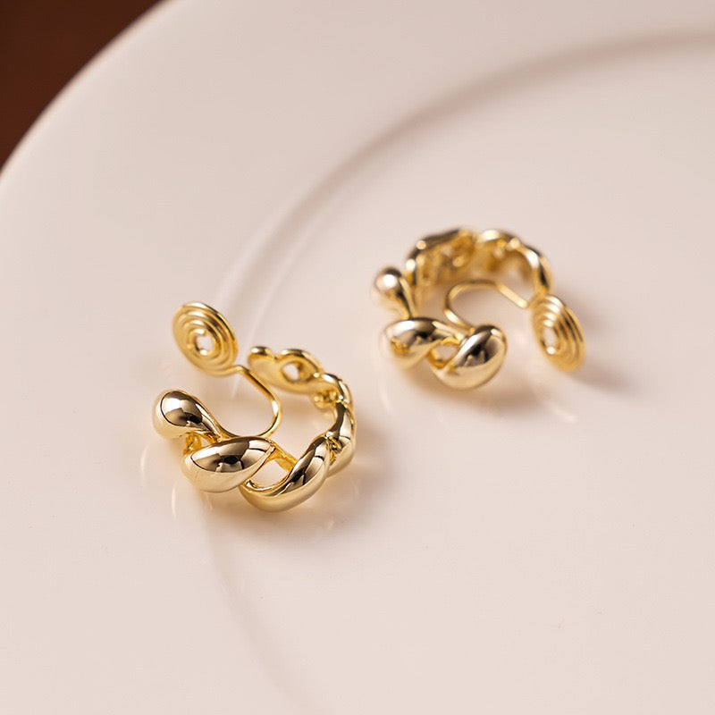 A pair of Earclipse gold twisted hoop clip-on earrings featuring a braided croissant design and spiral wire clip backings, resting on a white ceramic plate.