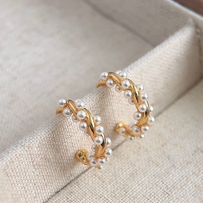 A pair of gold-tone hoop earrings featuring a twisted metal design intertwined with small pearls, shown with clear comfort clip-on backings on a white background.