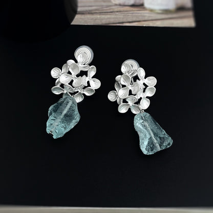 Pair of matte silver floral cluster earrings with dangling raw light blue crystals, highlighting the comfortable silicone-cushioned clip-on fittings on a dark background.