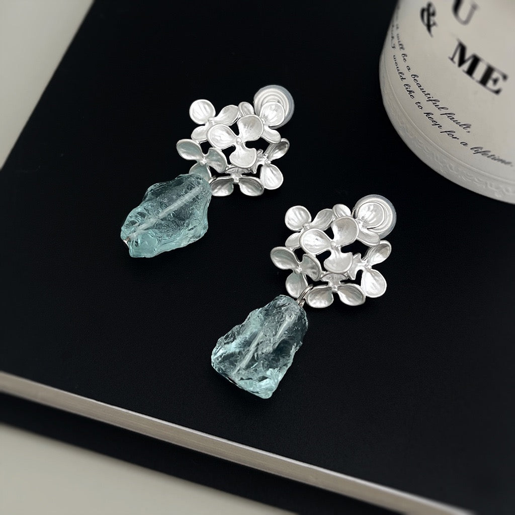 A pair of silver-toned dangle earrings with textured floral clusters and irregular light blue glass drops, displayed on a black book next to a white bottle.