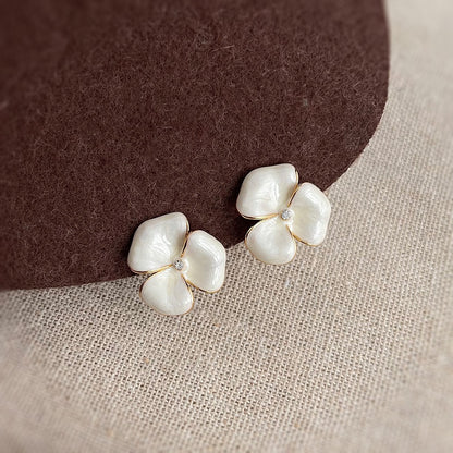 A pair of Earclipse pearlescent white four-petal flower stud earrings with gold-tone edging and a small clear crystal centre, resting on a textured brown and beige surface.