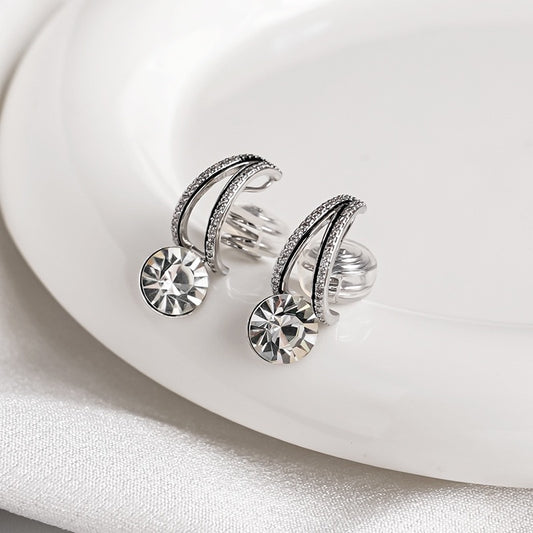 Pair of silver crystal solitaire drop clip-on earrings with pave-set double bands resting on a textured black surface and white silk