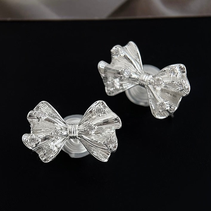 Silver textured ribbon bow clip-on earrings resting aesthetically on a soft surface next to an Earclipse branded velvet jewellery pouch