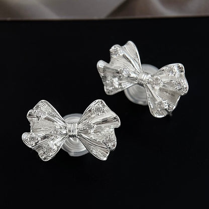 Silver textured ribbon bow clip-on earrings resting aesthetically on a soft surface next to an Earclipse branded velvet jewellery pouch