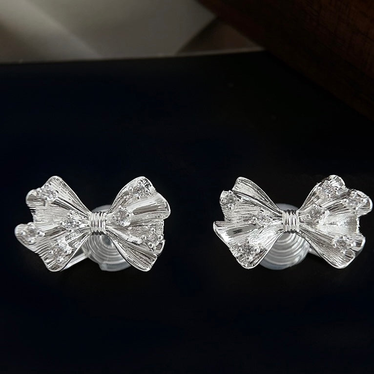 A pair of Earclipse silver clip-on stud earrings shaped like textured tied bows with small clear cubic zirconia crystals scattered across the ribbons, set against a white background