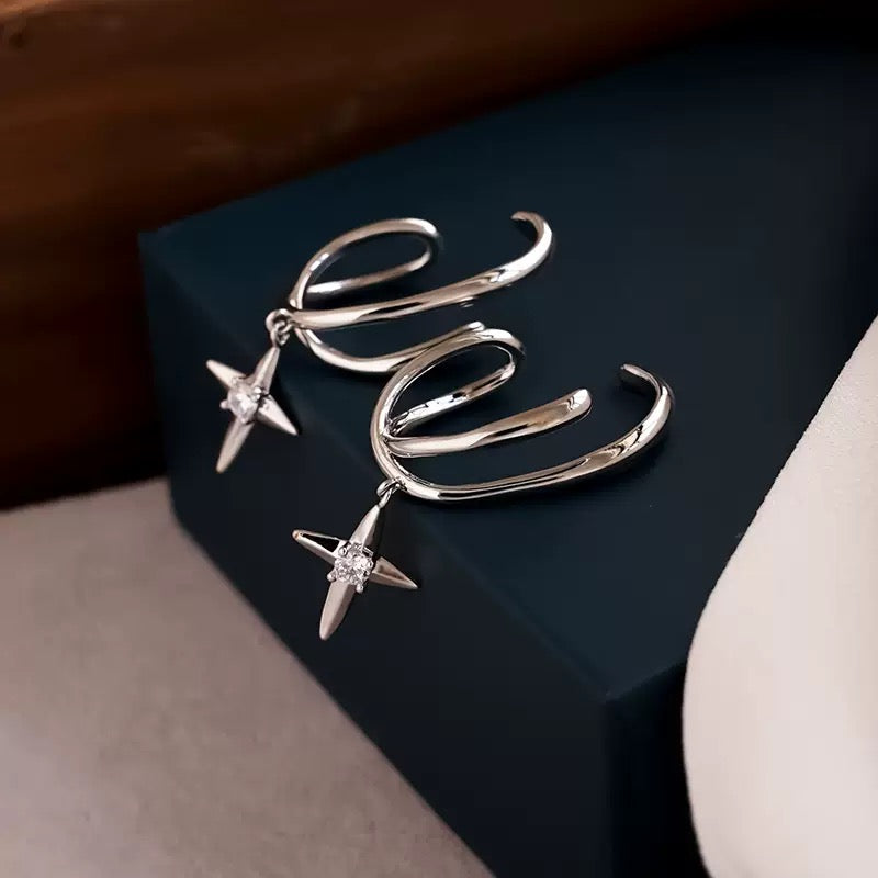 A pair of silver-toned statement earrings with abstract curved wire loops and dangling star-shaped charms set with clear stones, displayed on a dark blue jewelry box.